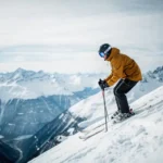 Skiër in okergeel jack aan de rand van steile Alpenhelling, met blik op besneeuwd dal en scherpe bergtoppen onder bewolkte winterlucht.