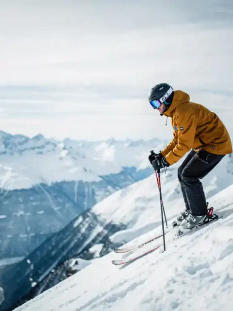 Skiër in okergeel jack aan de rand van steile Alpenhelling, met blik op besneeuwd dal en scherpe bergtoppen onder bewolkte winterlucht.