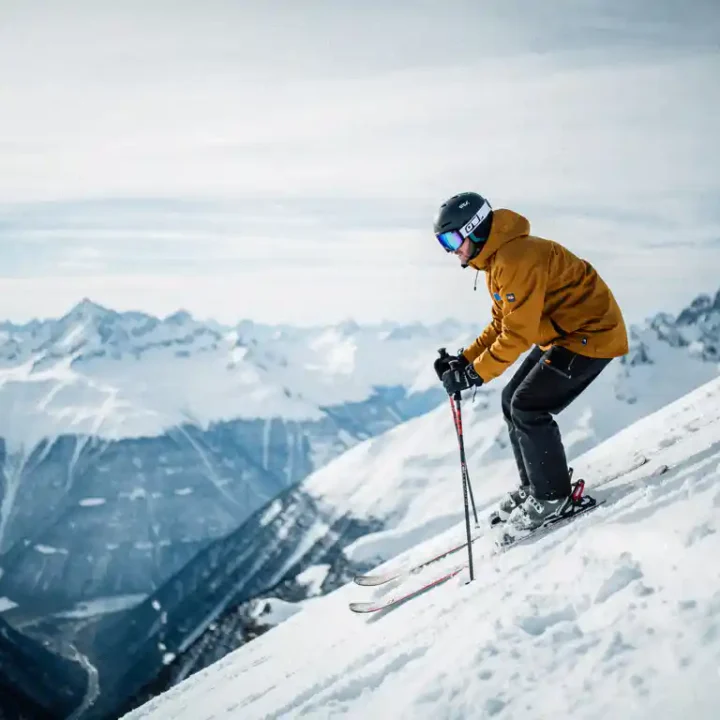 Skiër in okergeel jack aan de rand van steile Alpenhelling, met blik op besneeuwd dal en scherpe bergtoppen onder bewolkte winterlucht.