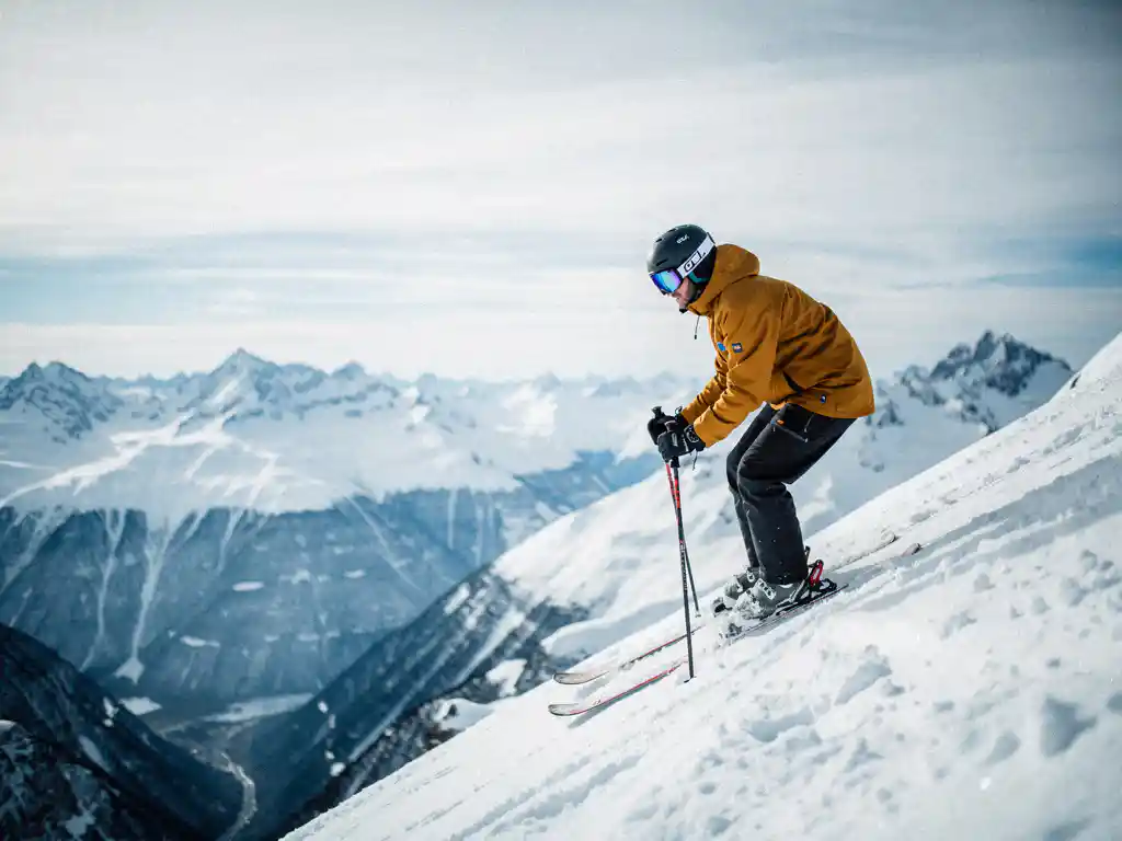 Skiër in okergeel jack aan de rand van steile Alpenhelling, met blik op besneeuwd dal en scherpe bergtoppen onder bewolkte winterlucht.