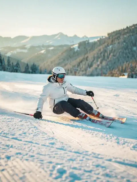 Skiër maakt een bocht op een verzorgde blauwe piste met glinsterende sneeuw, dennenbossen en heldere Alpiene lucht op de achtergrond.