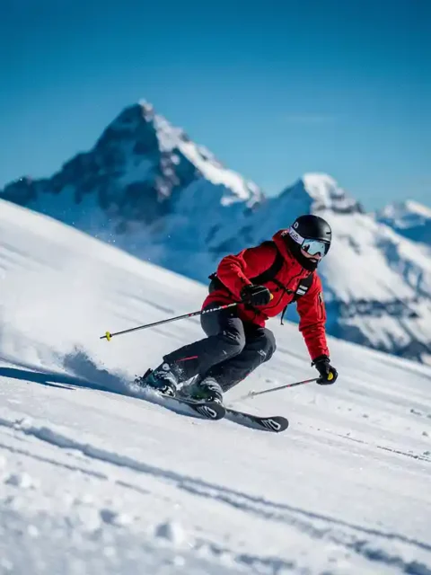 Skiër in perfecte carvingtechniek op verzorgde Alpijnse piste, rode jas, knieën gebogen, bergtoppen en blauwe lucht op achtergrond.