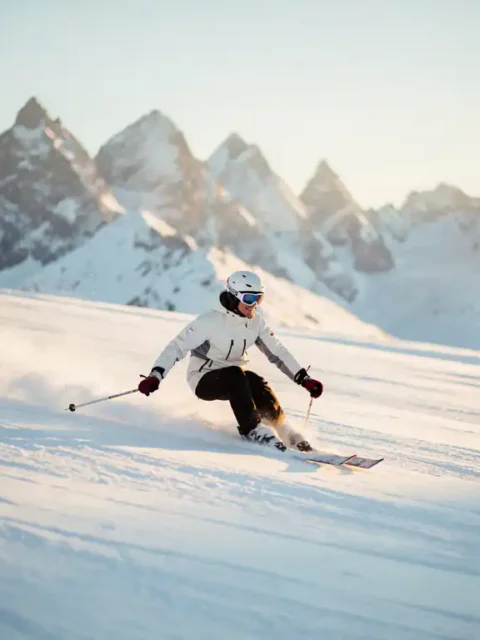 Skiër maakt een vloeiende bocht door poedersneeuw op een Alpiene piste, met bergtoppen en gouden ochtendzon op de achtergrond.