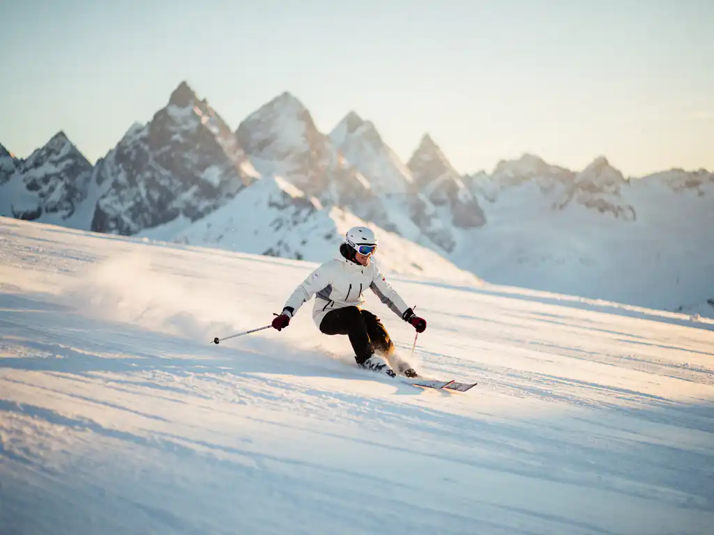 Skiër maakt een vloeiende bocht door poedersneeuw op een Alpiene piste, met bergtoppen en gouden ochtendzon op de achtergrond.