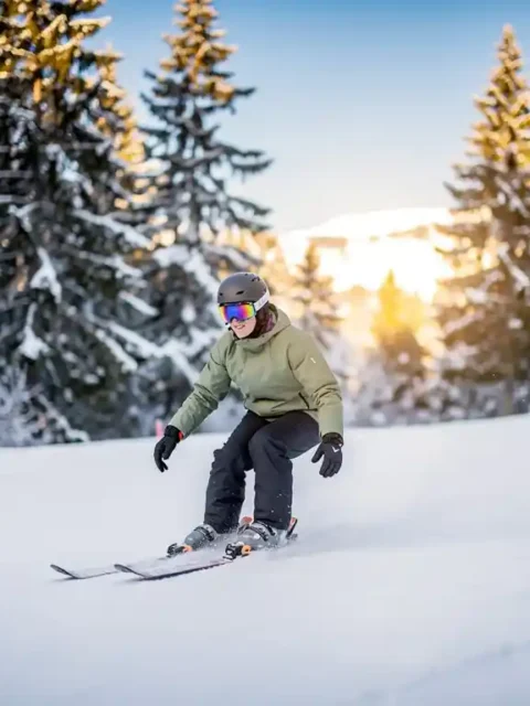 Beginnende skiër glijdt op een brede, zachte helling met besneeuwde pijnbomen en gouden alpiene zonlicht op de achtergrond.