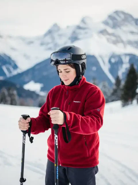 Beginnende skiër op besneeuwde helling houdt skistok vast en bestudeert deze, met beboste Alpiene toppen op de achtergrond.
