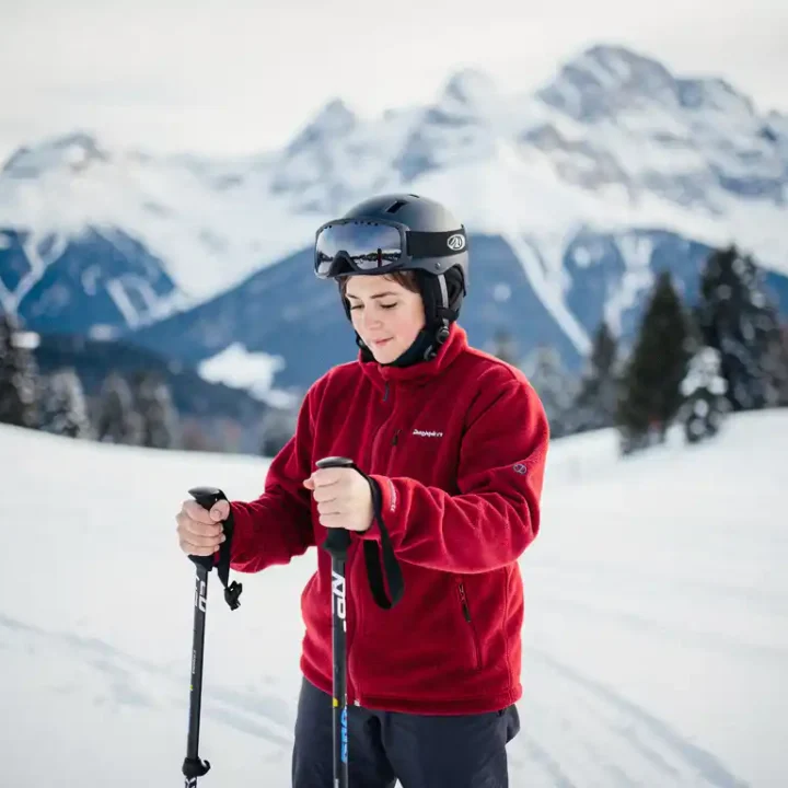 Beginnende skiër op besneeuwde helling houdt skistok vast en bestudeert deze, met beboste Alpiene toppen op de achtergrond.