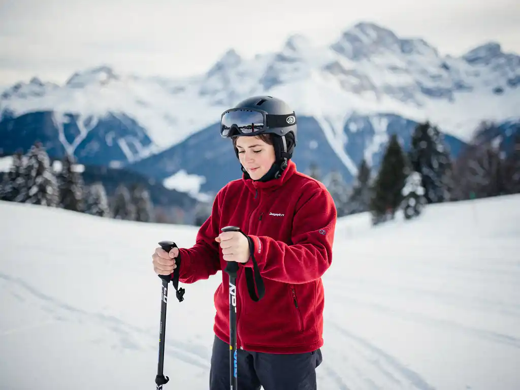Beginnende skiër op besneeuwde helling houdt skistok vast en bestudeert deze, met beboste Alpiene toppen op de achtergrond.
