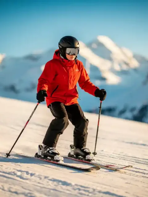 Beginnende skiër in rode jas maakt pizzawig op zachte besneeuwde helling, met besneeuwde Alpentoppen op de achtergrond.