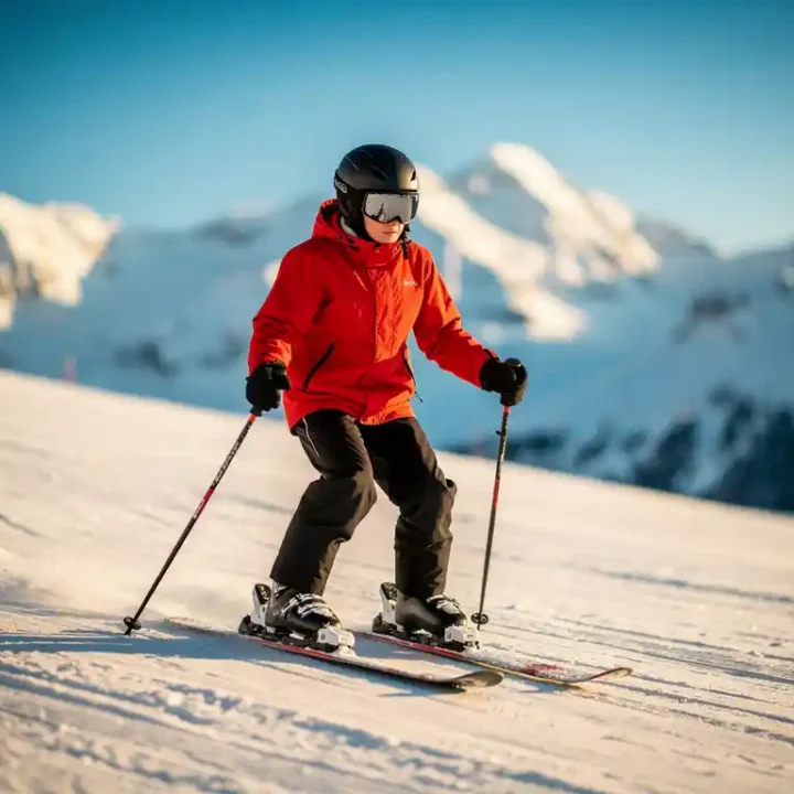 Beginnende skiër in rode jas maakt pizzawig op zachte besneeuwde helling, met besneeuwde Alpentoppen op de achtergrond.