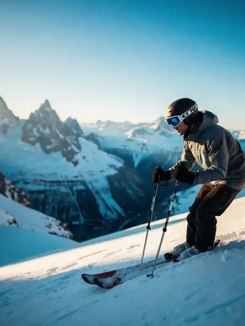 Skiër in volledige uitrusting aan de rand van een steile Alpenhelling, met uitzicht op een besneeuwd dal en scherpe bergtoppen onder een heldere blauwe lucht.