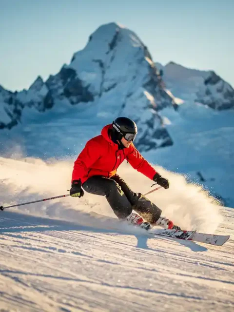 Skiër maakt een scherpe bocht op een verzorgde alpiene piste, knieën diep gebogen, poedersneeuw spat van de ski's onder stralende middagzon.