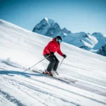 Skiër in rood jack maakt vloeiende bochten op verzorgde beginnersskihelling met Alpiene bergtoppen en blauwe lucht op de achtergrond.