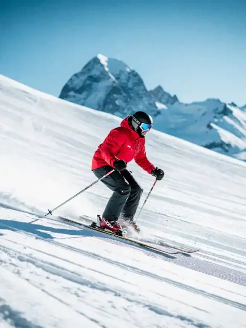 Skiër in rood jack maakt vloeiende bochten op verzorgde beginnersskihelling met Alpiene bergtoppen en blauwe lucht op de achtergrond.