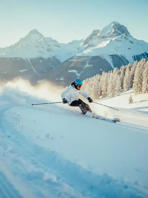 Beginner skiër maakt een brede bocht op een verzorgde Alpijnse piste met besneeuwde bergtoppen op de achtergrond.