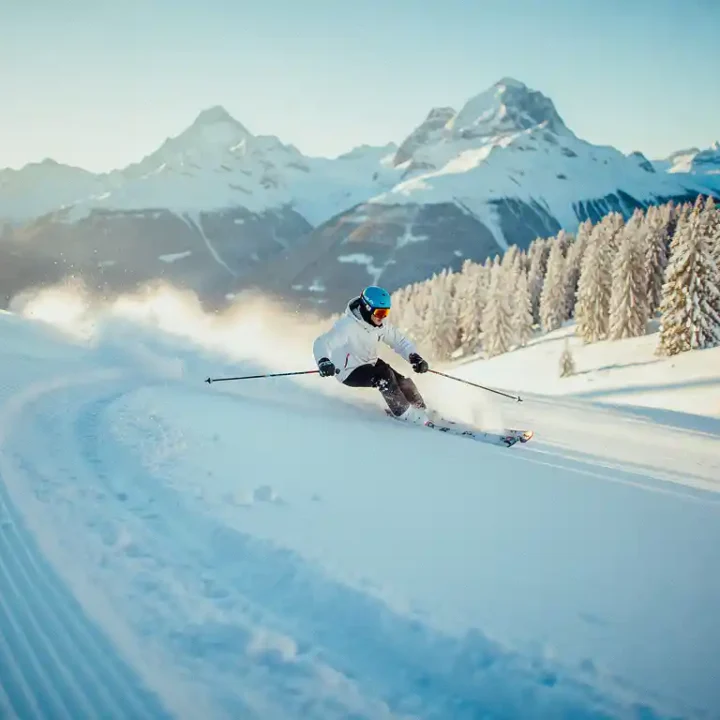 Beginner skiër maakt een brede bocht op een verzorgde Alpijnse piste met besneeuwde bergtoppen op de achtergrond.