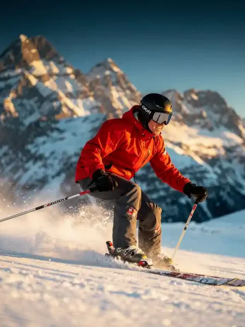 Skiër in rode jas snijdt een scherpe bocht op verzorgde Alpine piste, met bergtoppen en blauwe lucht op de achtergrond.