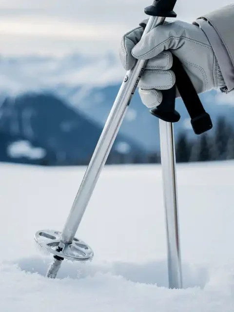 Gehandschoende hand met aluminium skistokken in verse sneeuw op een Alpijn skigebied, met besneeuwde bergtoppen op de achtergrond.