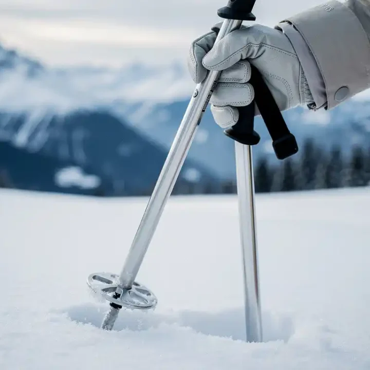 Gehandschoende hand met aluminium skistokken in verse sneeuw op een Alpijn skigebied, met besneeuwde bergtoppen op de achtergrond.
