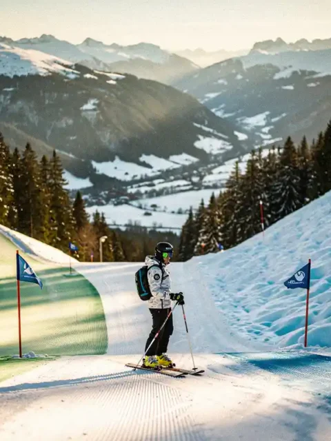 Skiër bij een kruispunt van groene en blauwe piste, met vers poedersneeuw en een Alpien dal omgeven door dennenbossen op de achtergrond.