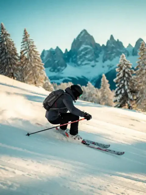 Skiër maakt een brede parallelle bocht op een verzorgde Alpine piste, omgeven door besneeuwde dennenbomen en bergtoppen in gouden zonlicht.