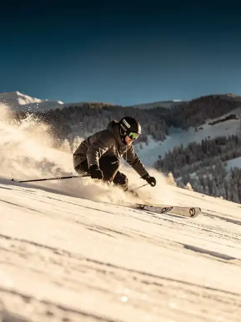 Skiër snijdt een bocht door verse poedersneeuw op steile Alpenhelling, sneeuwspray waaiert uit onder stralend blauwe lucht.