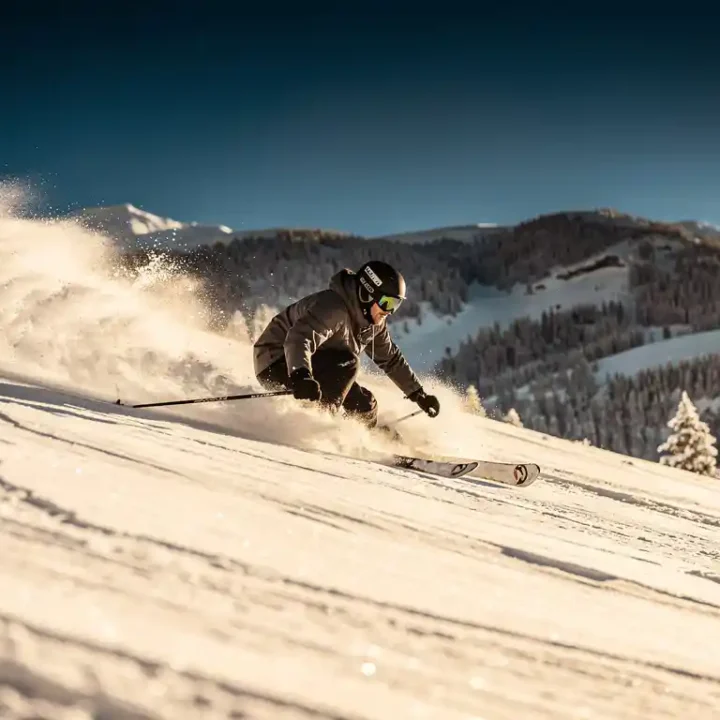 Skiër snijdt een bocht door verse poedersneeuw op steile Alpenhelling, sneeuwspray waaiert uit onder stralend blauwe lucht.