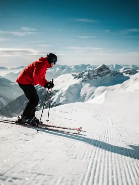 Skiër in rood jack staat boven steile, verzorgde alpine piste, stokken in de sneeuw, met uitzicht op besneeuwd bergpanorama.