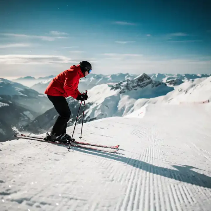 Skiër in rood jack staat boven steile, verzorgde alpine piste, stokken in de sneeuw, met uitzicht op besneeuwd bergpanorama.