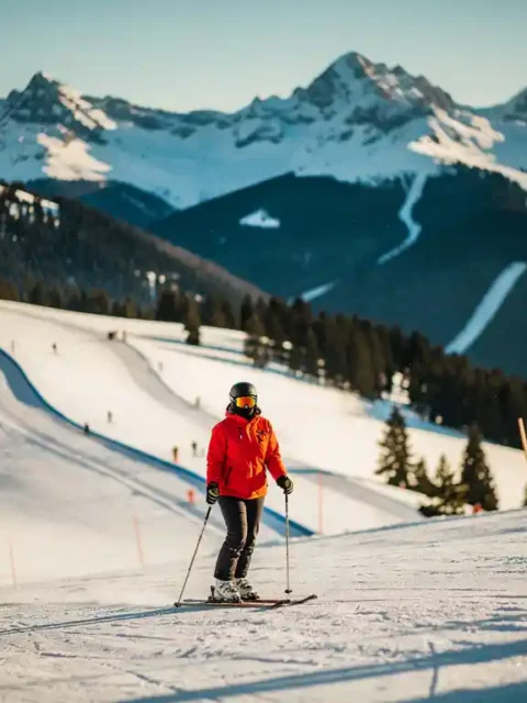 Skiër in felrood jack op een groene beginnersnpiste, met een steilere blauwe run en besneeuwde Alpiene toppen op de achtergrond.