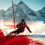 Skiër maakt een bocht op een verzorgde rode piste met Alpiene bergtoppen en strakblauwe lucht op de achtergrond.