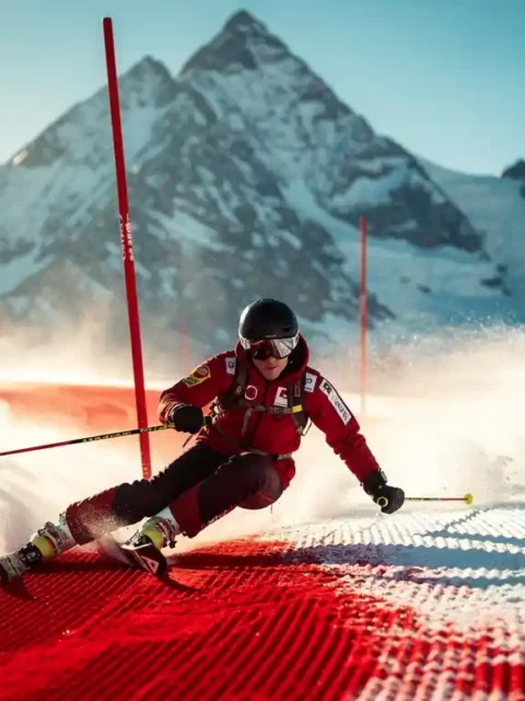 Skiër maakt een bocht op een verzorgde rode piste met Alpiene bergtoppen en strakblauwe lucht op de achtergrond.
