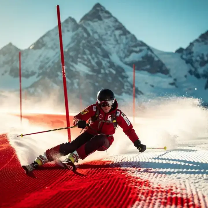 Skiër maakt een bocht op een verzorgde rode piste met Alpiene bergtoppen en strakblauwe lucht op de achtergrond.