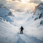 Skiër bovenaan een steile zwarte piste in de Alpen, uitkijkend over een besneeuwde couloir met scherpe bergtoppen op de achtergrond.