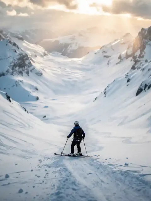 Skiër bovenaan een steile zwarte piste in de Alpen, uitkijkend over een besneeuwde couloir met scherpe bergtoppen op de achtergrond.