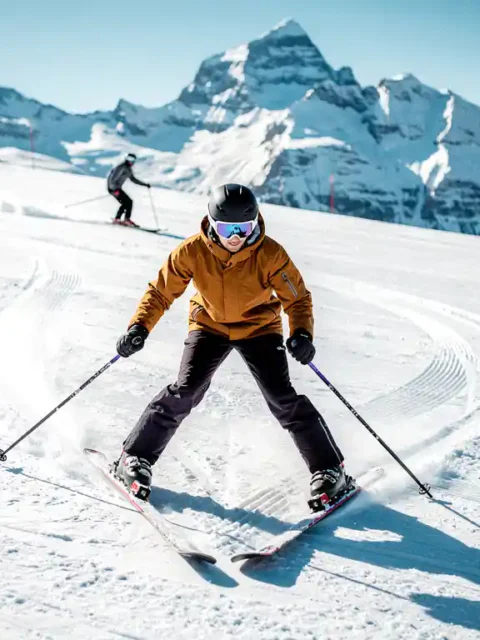 Ski-instructeur demonstreert sneeuwploeghouding met V-vormige ski's op verzorgde beginnersbaan, besneeuwde Alpentopen op de achtergrond.