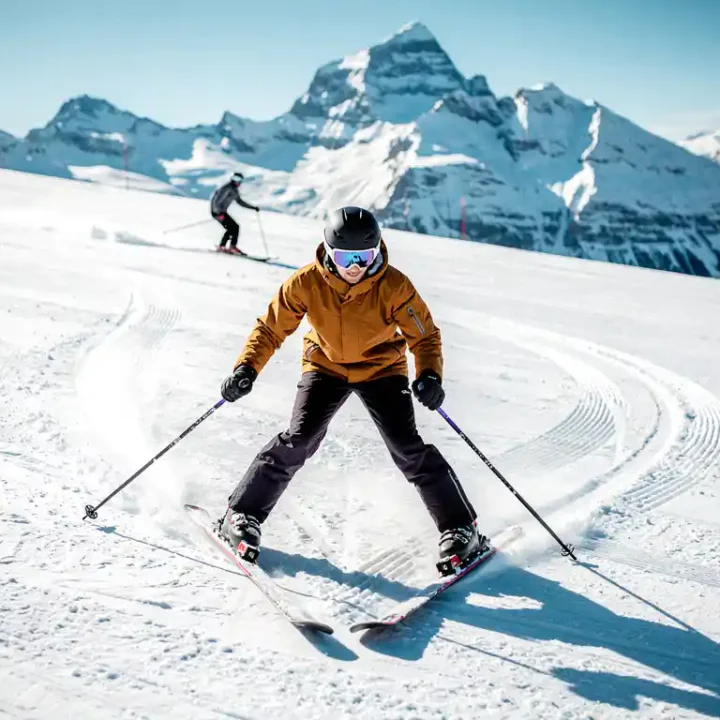Ski-instructeur demonstreert sneeuwploeghouding met V-vormige ski's op verzorgde beginnersbaan, besneeuwde Alpentopen op de achtergrond.