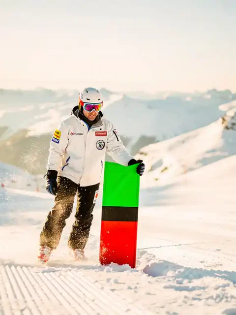 Skipatrouillewerker plaatst groene, rode en zwarte pistemarcering in verse sneeuw op een verzorgde Alpijnse piste bij goudkleurig ochtendlicht.
