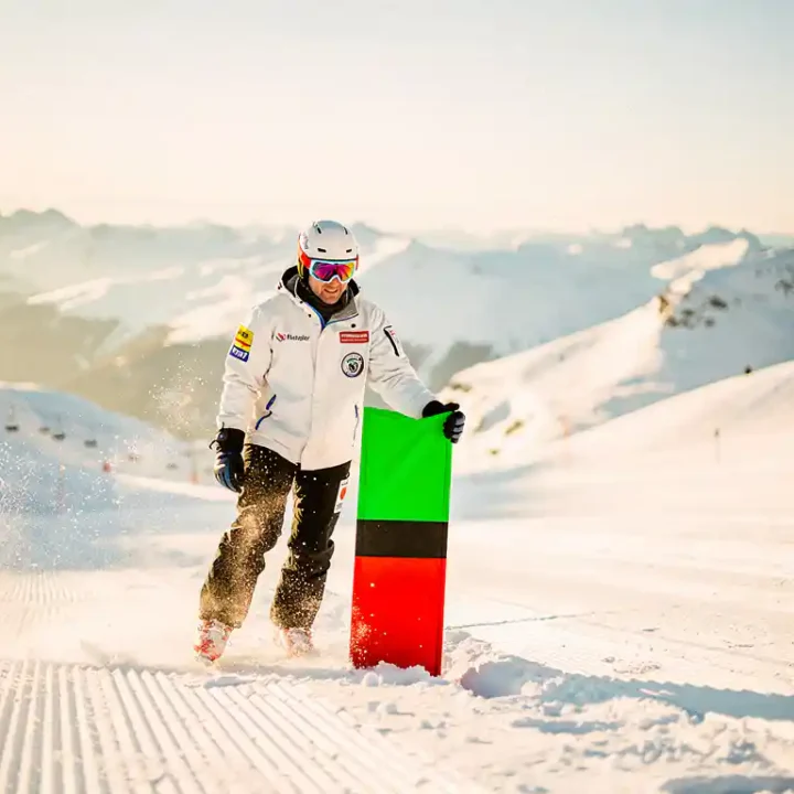 Skipatrouillewerker plaatst groene, rode en zwarte pistemarcering in verse sneeuw op een verzorgde Alpijnse piste bij goudkleurig ochtendlicht.