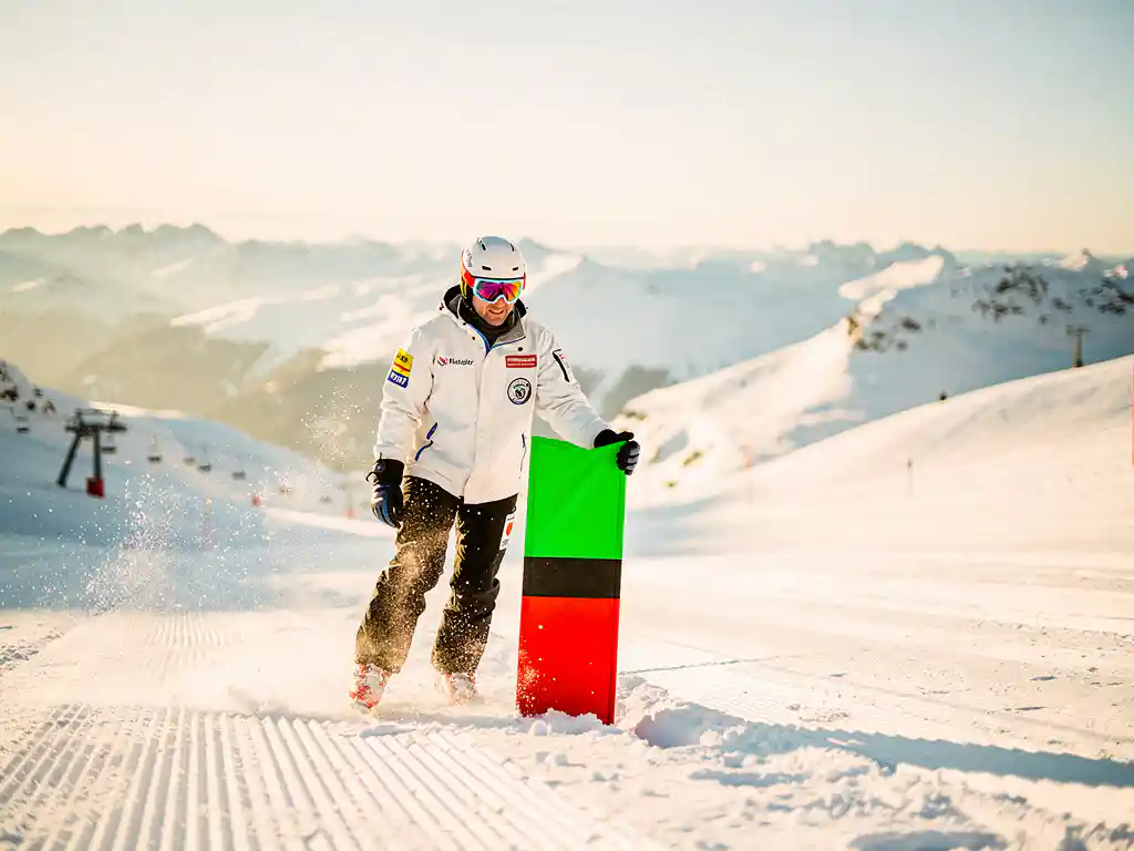 Skipatrouillewerker plaatst groene, rode en zwarte pistemarcering in verse sneeuw op een verzorgde Alpijnse piste bij goudkleurig ochtendlicht.