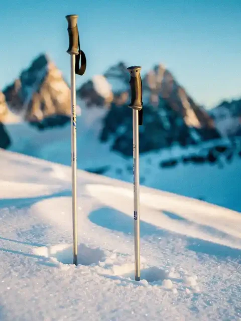 Twee skipoles rechtop in poedersneeuw op een alpiene helling, met dramatische bergtoppen en blauwe lucht op de achtergrond.