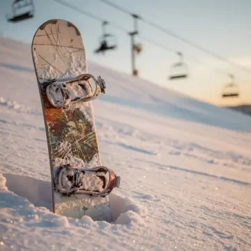 Gebrauchtes Snowboard mit Gebrauchsspuren lehnt an Pulverschnee auf Bergpiste, Skilift im Hintergrund, goldenes Licht