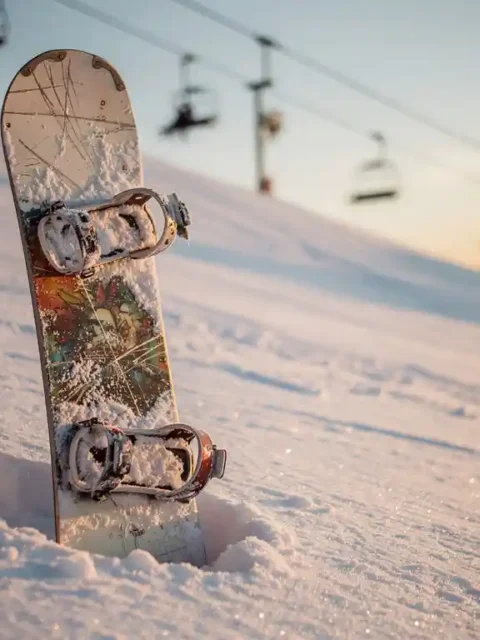 Gebrauchtes Snowboard mit Gebrauchsspuren lehnt an Pulverschnee auf Bergpiste, Skilift im Hintergrund, goldenes Licht