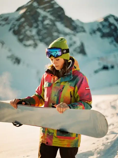 Beginnende snowboarder in felgekleurde winterkleding bekijkt snowboard op besneeuwde berghellingen tijdens gouden uur