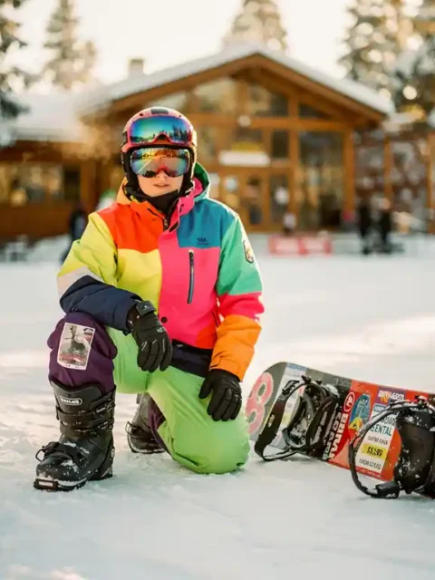 Beginnende snowboarder knielt naast huursnowboard op verse sneeuw, ski lodge op achtergrond, gouden zonlicht