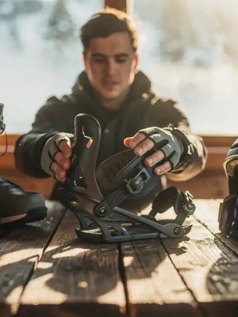 Snowboarder inspecteert snowboard binding aan houten hut tafel met laarzen, bril en helm in warme winterse sfeer