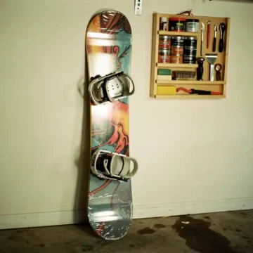 Snowboard lehnt an Garagenwand mit Schutzabdeckungen, Wandregal mit Wachsausrüstung und Tuning-Werkzeugen sichtbar