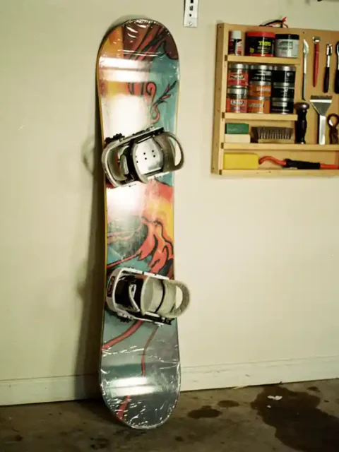 Snowboard lehnt an Garagenwand mit Schutzabdeckungen, Wandregal mit Wachsausrüstung und Tuning-Werkzeugen sichtbar