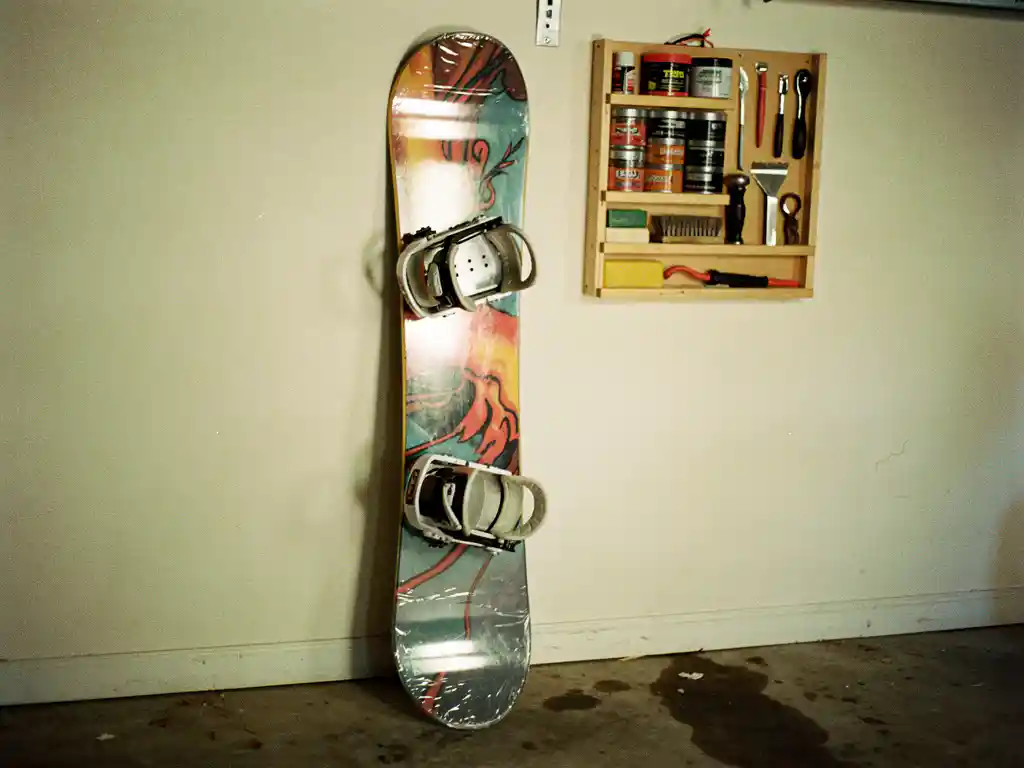 Snowboard lehnt an Garagenwand mit Schutzabdeckungen, Wandregal mit Wachsausrüstung und Tuning-Werkzeugen sichtbar