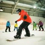 Snowboard instructeur in rode jas toont juiste houding aan beginners op kunstsneeuw in overdekte skifaciliteit